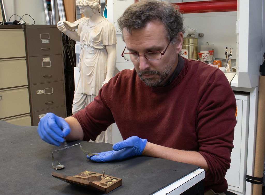 Man sat at a table holding a small brass balance used for weighing coins, with a small wooden box on the table in front