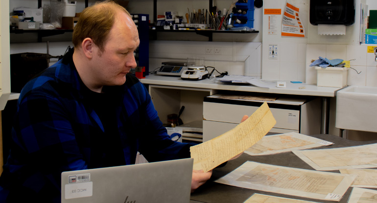 A man sat at a table with various documents, holding a document. Surrounded by conservation lab equipment