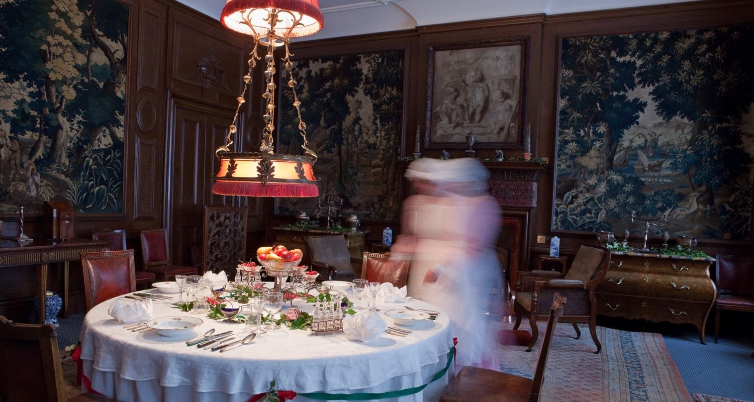 A ghostly image of a maid laying the large round dining table in the Lauriston Castle dining room. An ornate lamp hangs over the table and the walls are covered with tapestries 