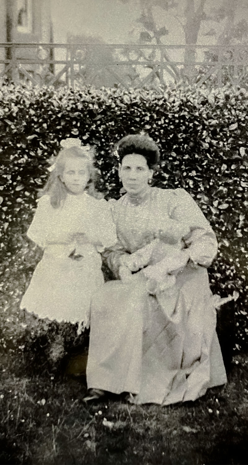 a faded and blurred sepia photograph of a young girl in a white dress with a woman. The woman is holding a teddy bear.