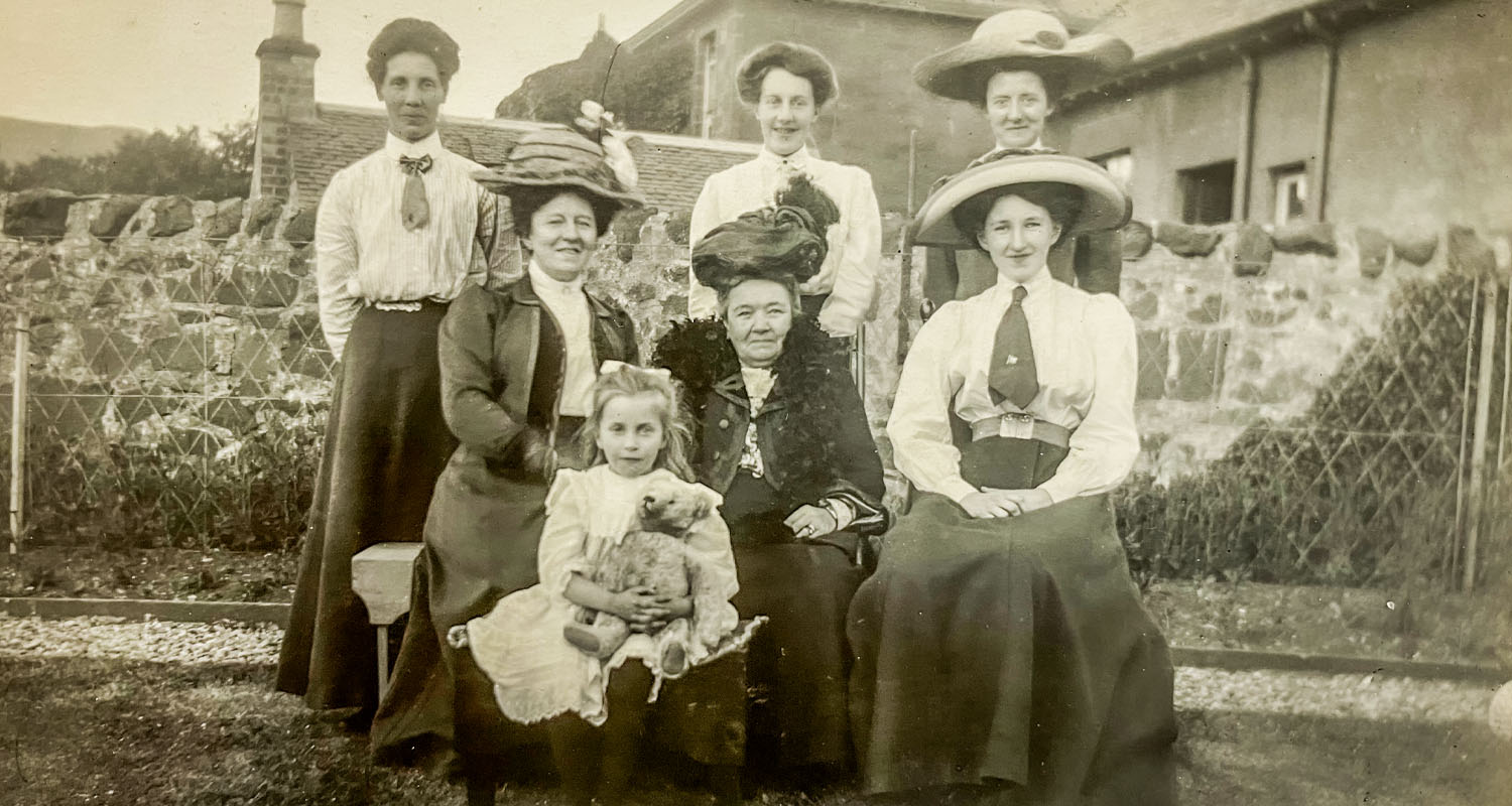 Sepia photograph of a group of women in Edwardian costume with a young girl at the front holding a teddy bear. Behind them is a large farmhouse building.