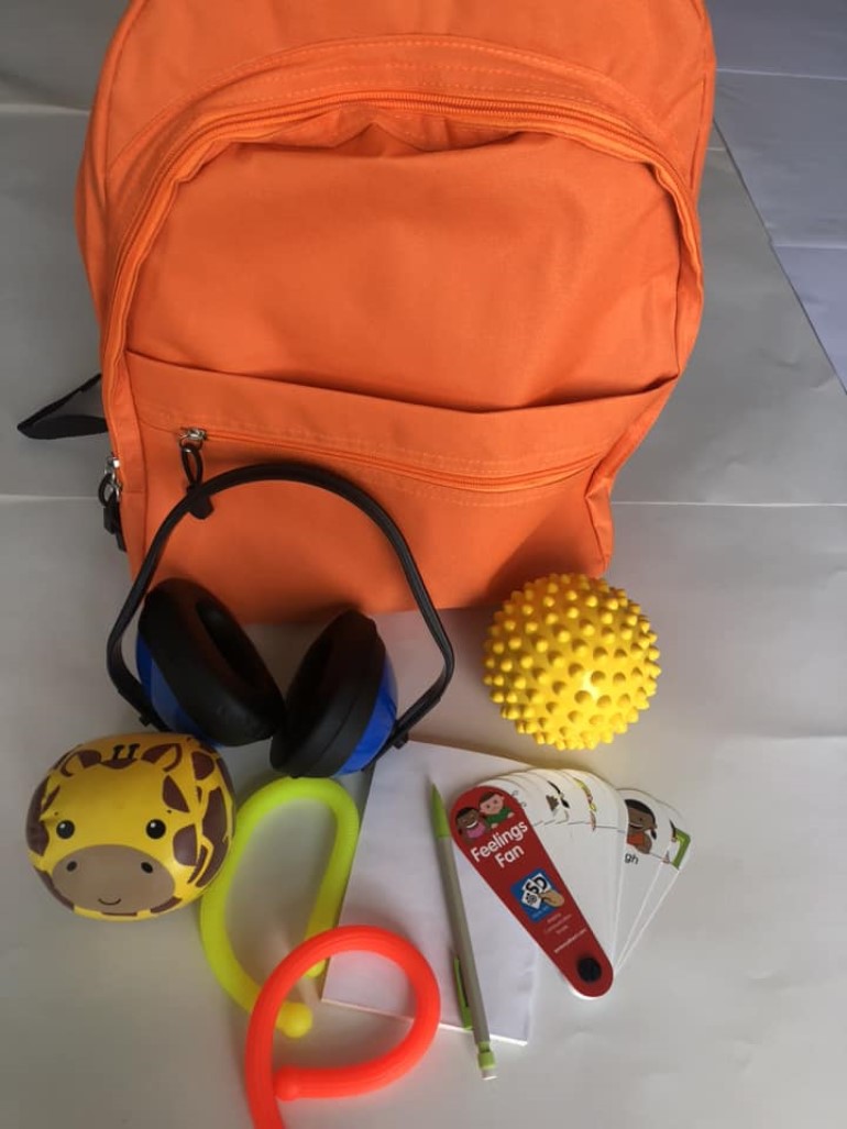 An orange backpack with various sensory toys, including ear defenders, fidget toys, a notepad and pencil and emotion cards