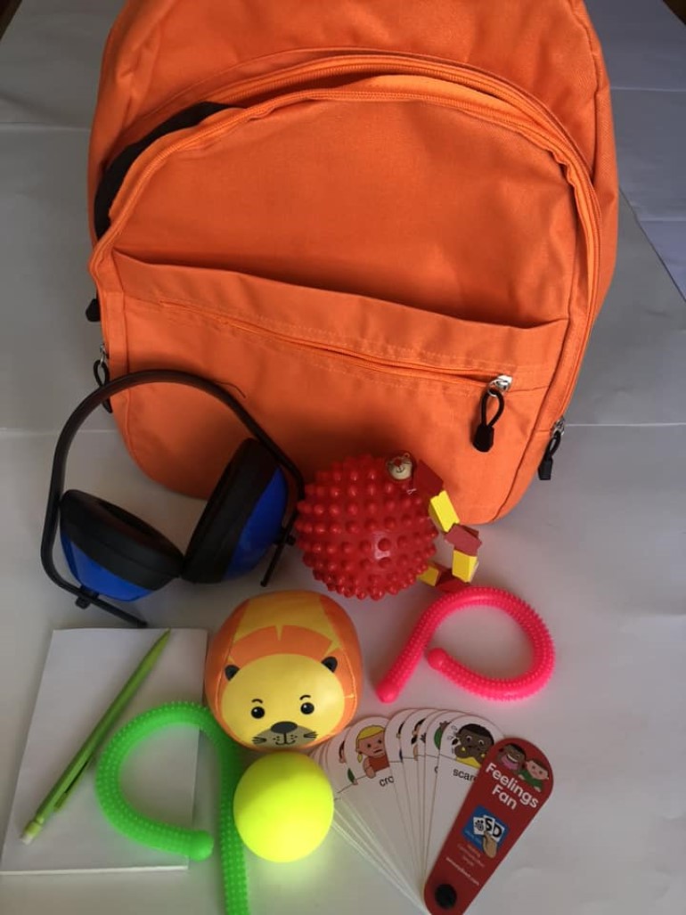 An orange backpack with various sensory toys, including ear defenders, fidget toys, a notepad and pencil and emotion cards