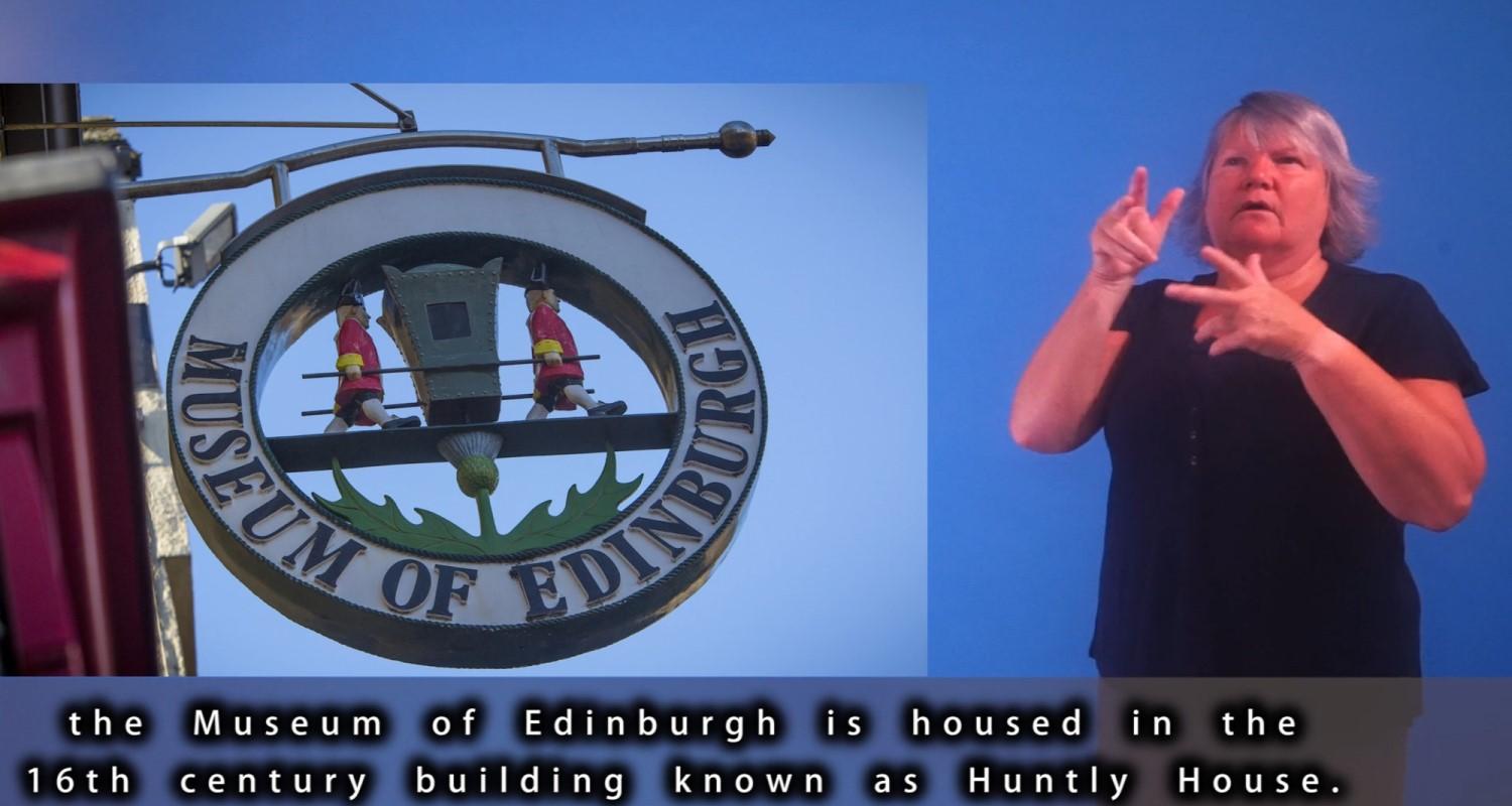 A BSL interpreter beside a photo of the Museum of Edinburgh sign. Subtitles running along the bottom mention that the museum is housed in 16th century building Huntly House