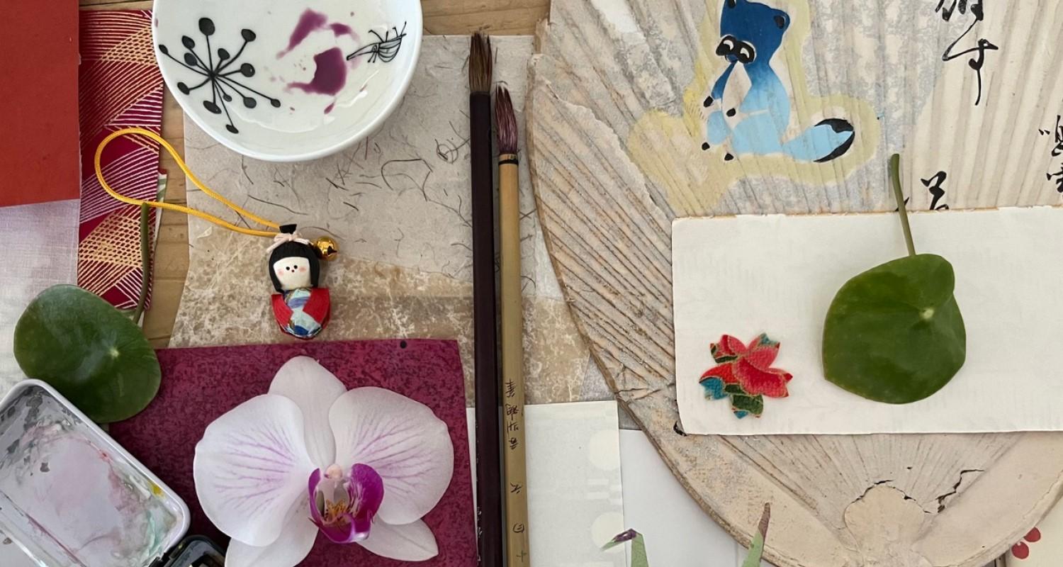 A tabletop covered in paintbrushes, a paintbox, and Japanese miscellany including a fan and a small doll