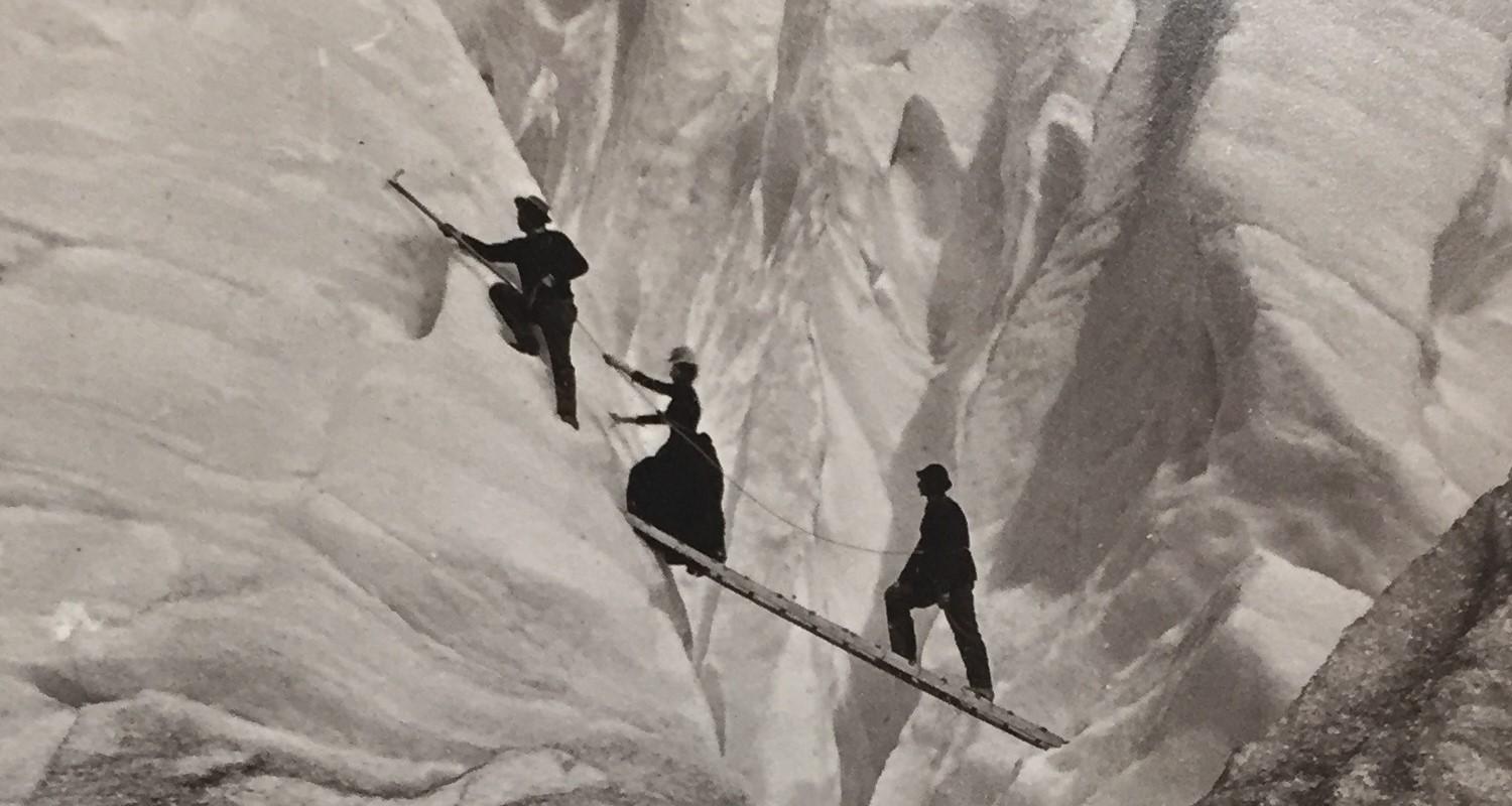 Two men and a woman crossing a snowy crevasse on a ladder. The men are tied together by a rope, the one in front has a pickaxe embedded in the snow. The woman in the middle wears a hat and a long dress(!)