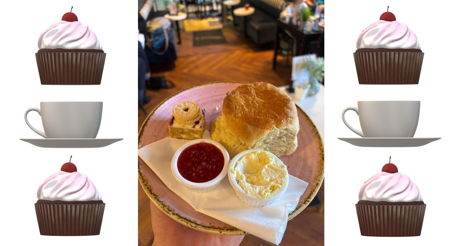 A plate held above a cafe floor containing a plan scone, and pots of jam and cream with a biscuit and a traybake