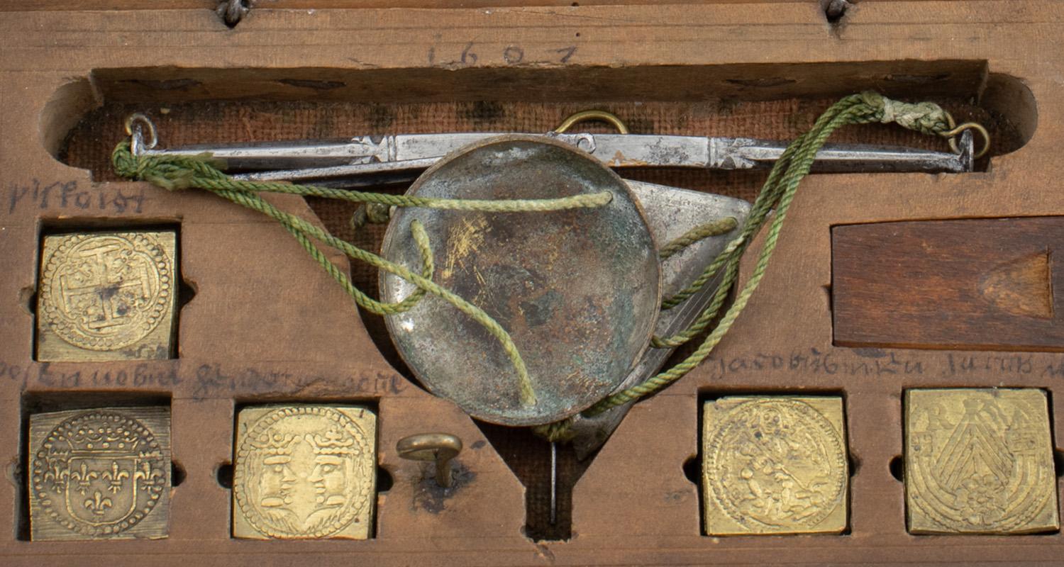 Close up of a wooden box with embossed brass square coin weights and a small weighing scale with metal pans