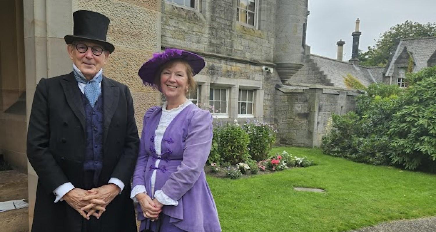 A male ELH performer in a top hat and suit and female performer in a lilac dress and hat stand in front of Lauriston Castle