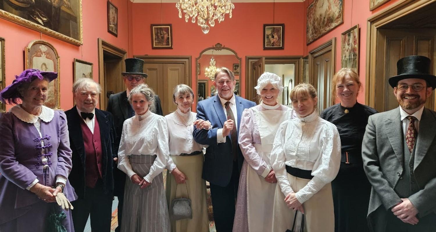 A group of people in Edwardian dress of staff and residents stand in a passageway in the castle beneath a chandelier. The walls are covered in artworks; five doors lead off