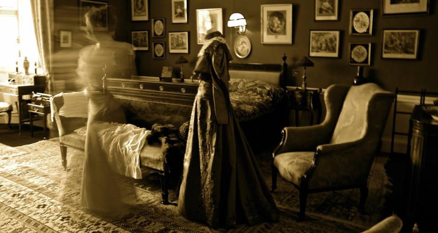 A sepia image of two ghostly images of women in Edwardian dress in a living room full of furniture with paintings on the wall