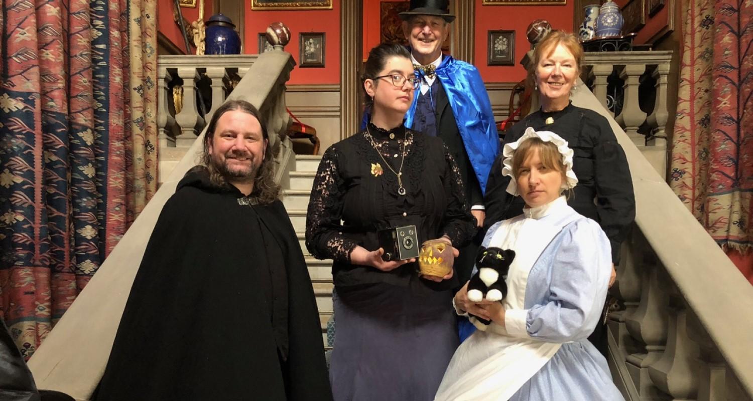 Lauriston Castle stff standing on the staircase, some in cloaks, some in uniform. One holds a turnip lantern, the other a soft toy cat.