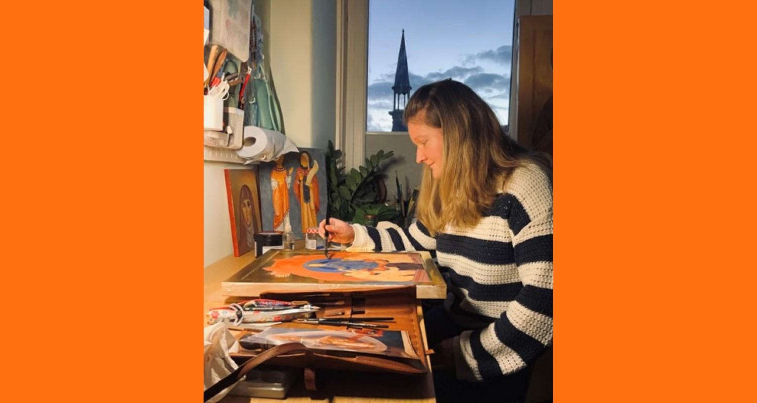 A woman in a black and white striped jumper sitting in front of a window painting icons