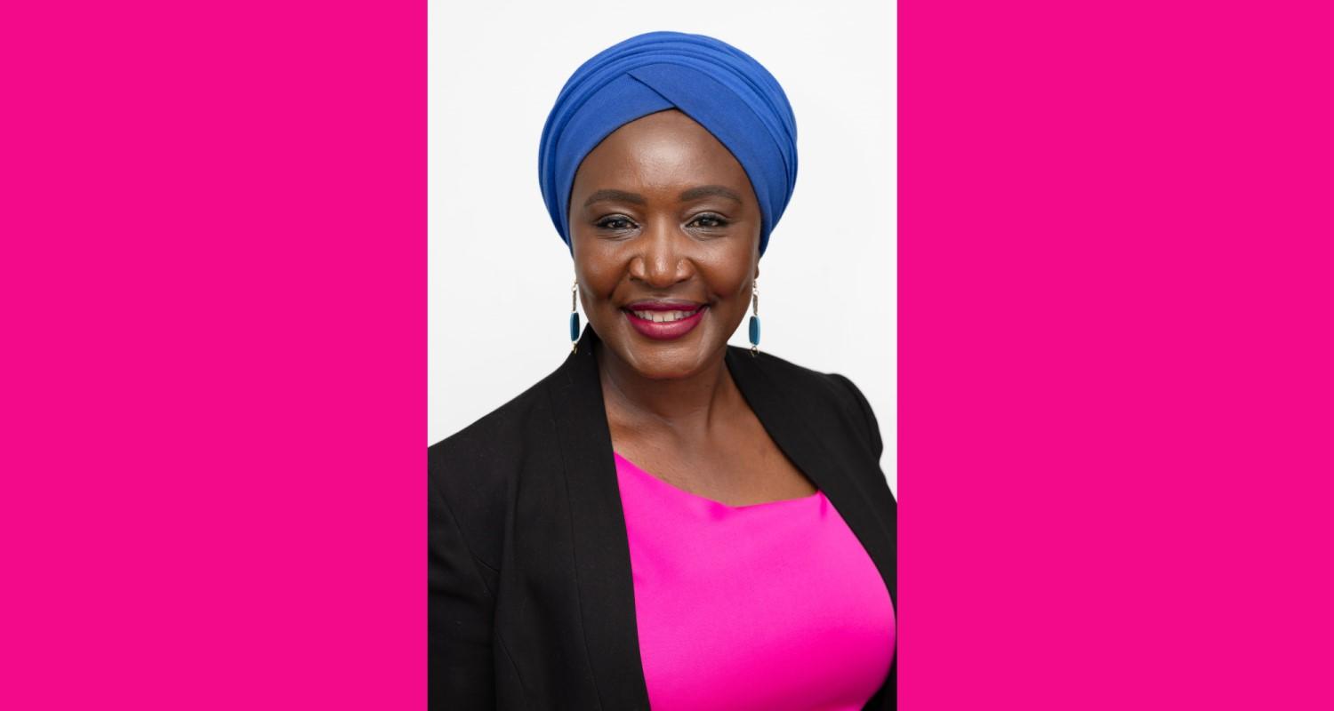 A head and shoulders portrait of Irene Mosota, wearing a pink top, black jacket and blue head wrap