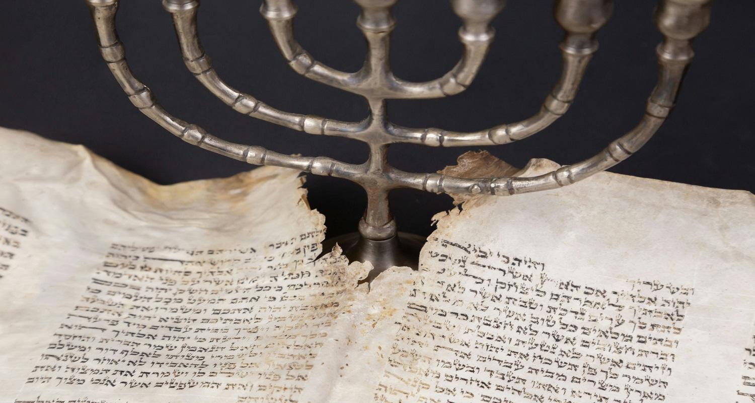 A silver menorah between a ripped, burned page of Hebrew text