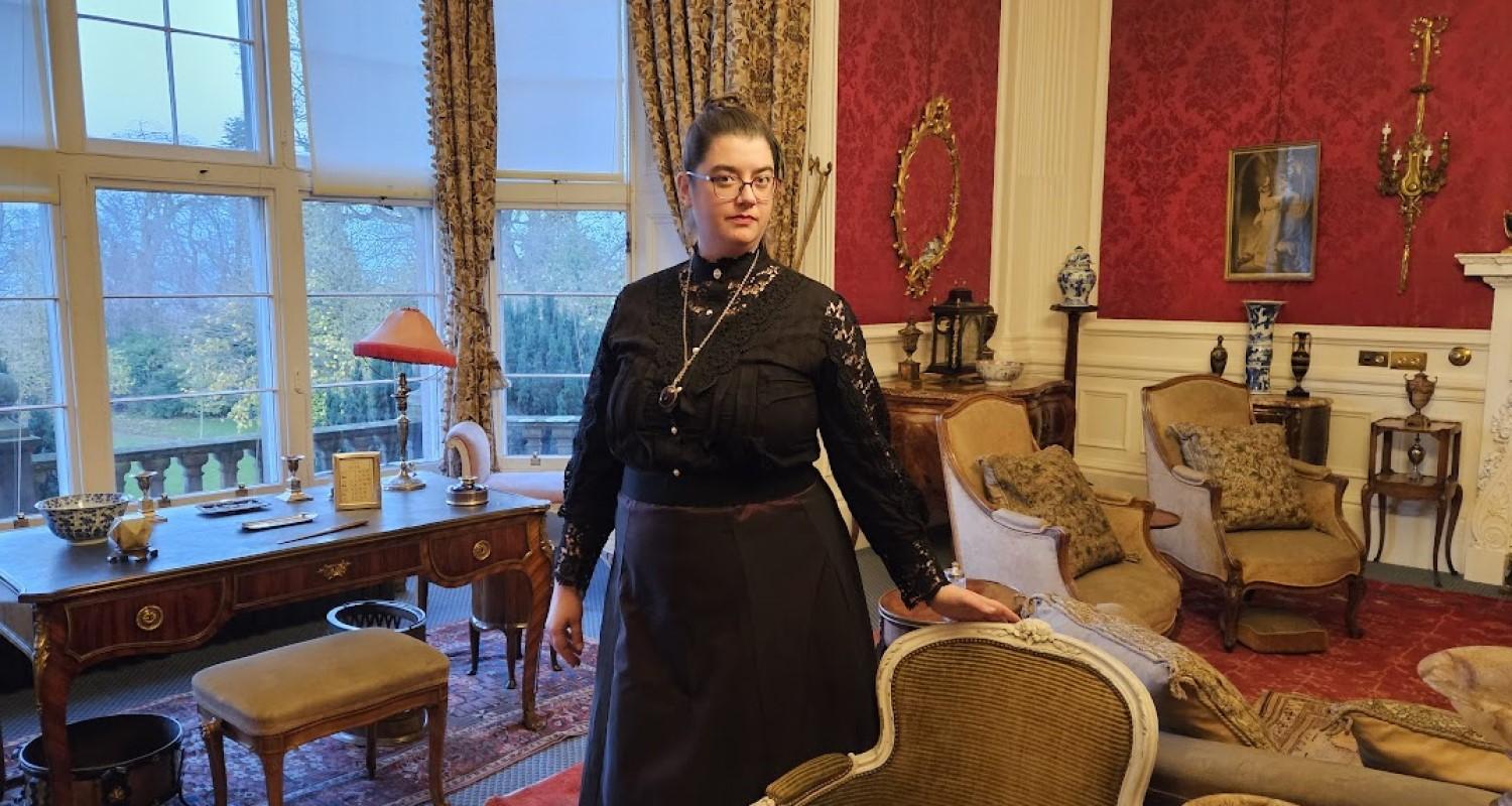 An Edinburgh Living History performer wearing a black dress standing in the living room at Lauriston Castle. Surrounded by chairs and a desk, outside the window the garden and trees can be seen