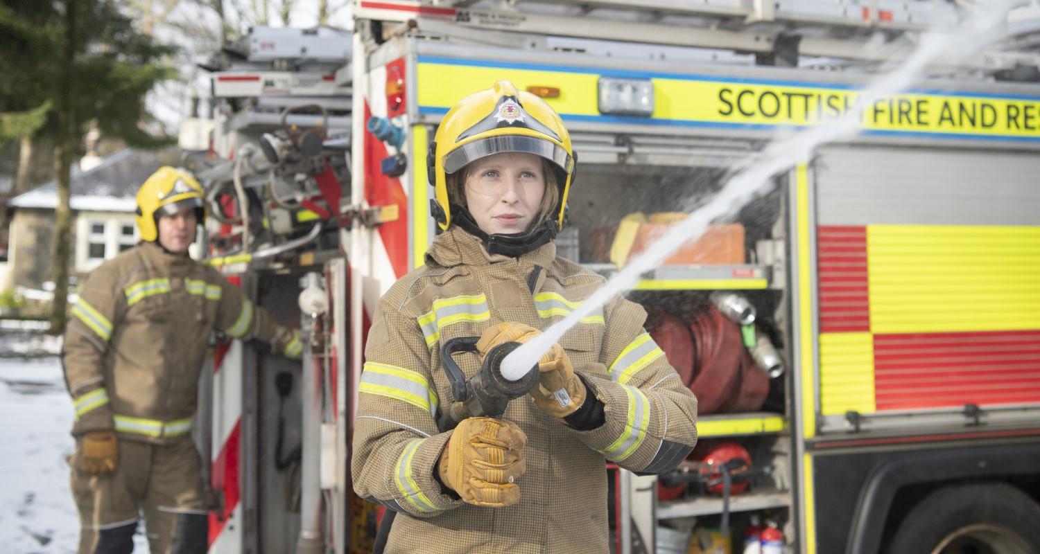 A firefighter in uniform stands in front of a fire engine, hosing an object off-camera