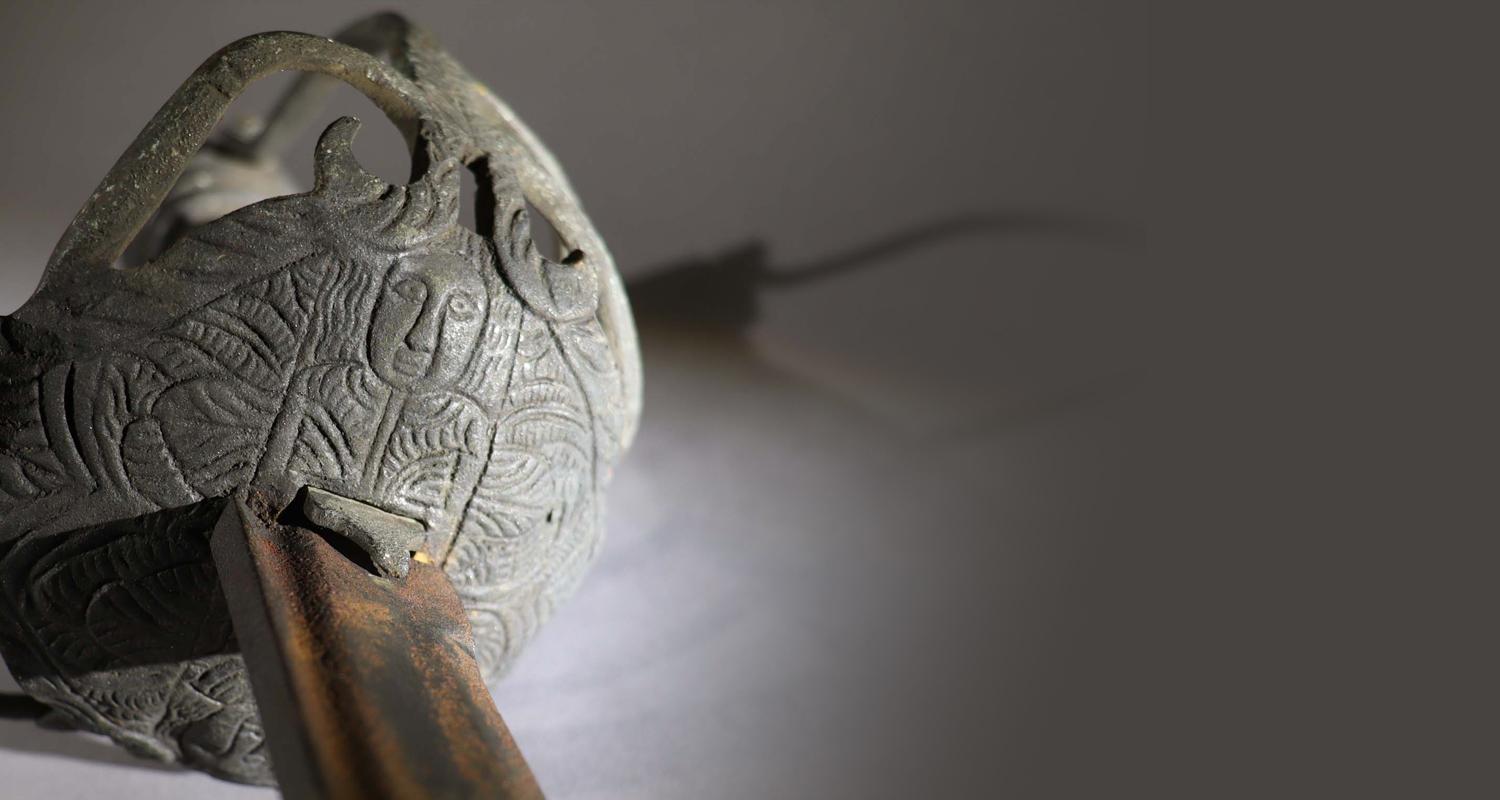 A close up of a sword hilt and blade of the type known as a mortuary, with an engraved stylised head.