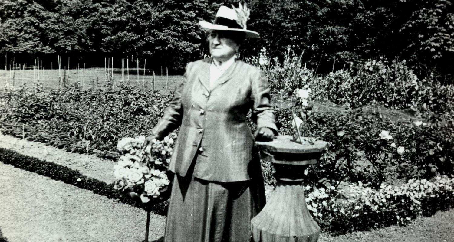 Mrs Reid in a long skirt and jacket, wearing a hat and carrying a cane, standing beside a sundial in the garden. Black and white photo