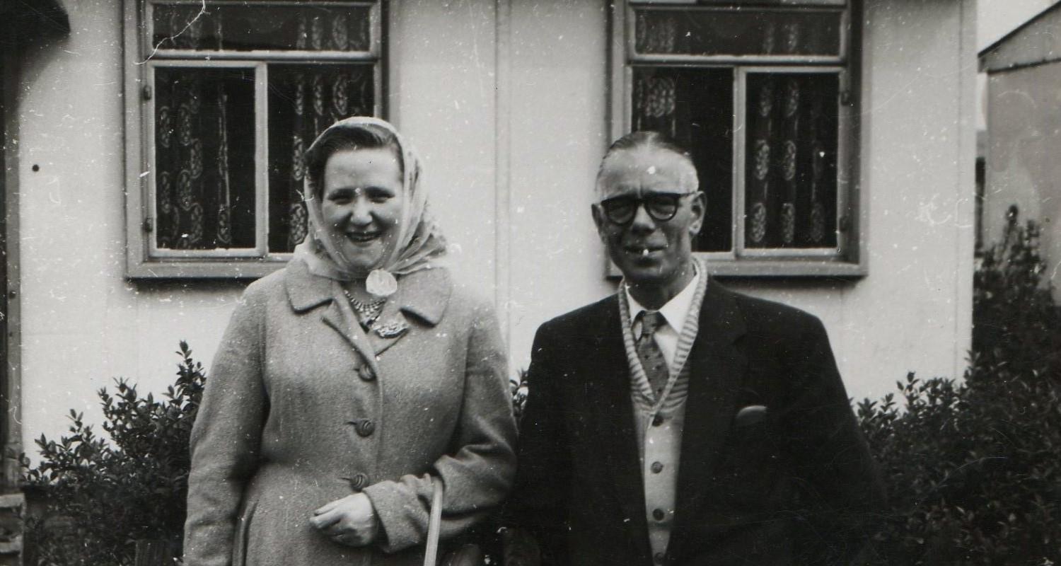 A black and white photo of a middle aged couple standing outside a house. She wears a winter coat and headscarf and has a handbag looped over her arm, he wears a coat over a cardigan, shirt and tie. There is a cigarette in his mouth.