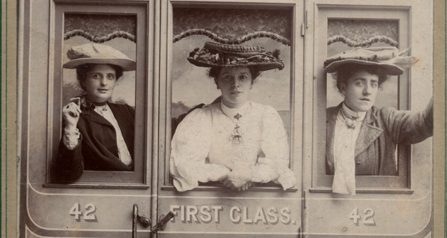 A black and white photo of three women in Edwardian clothes and big hats leaning out of a train window from the first class carriage