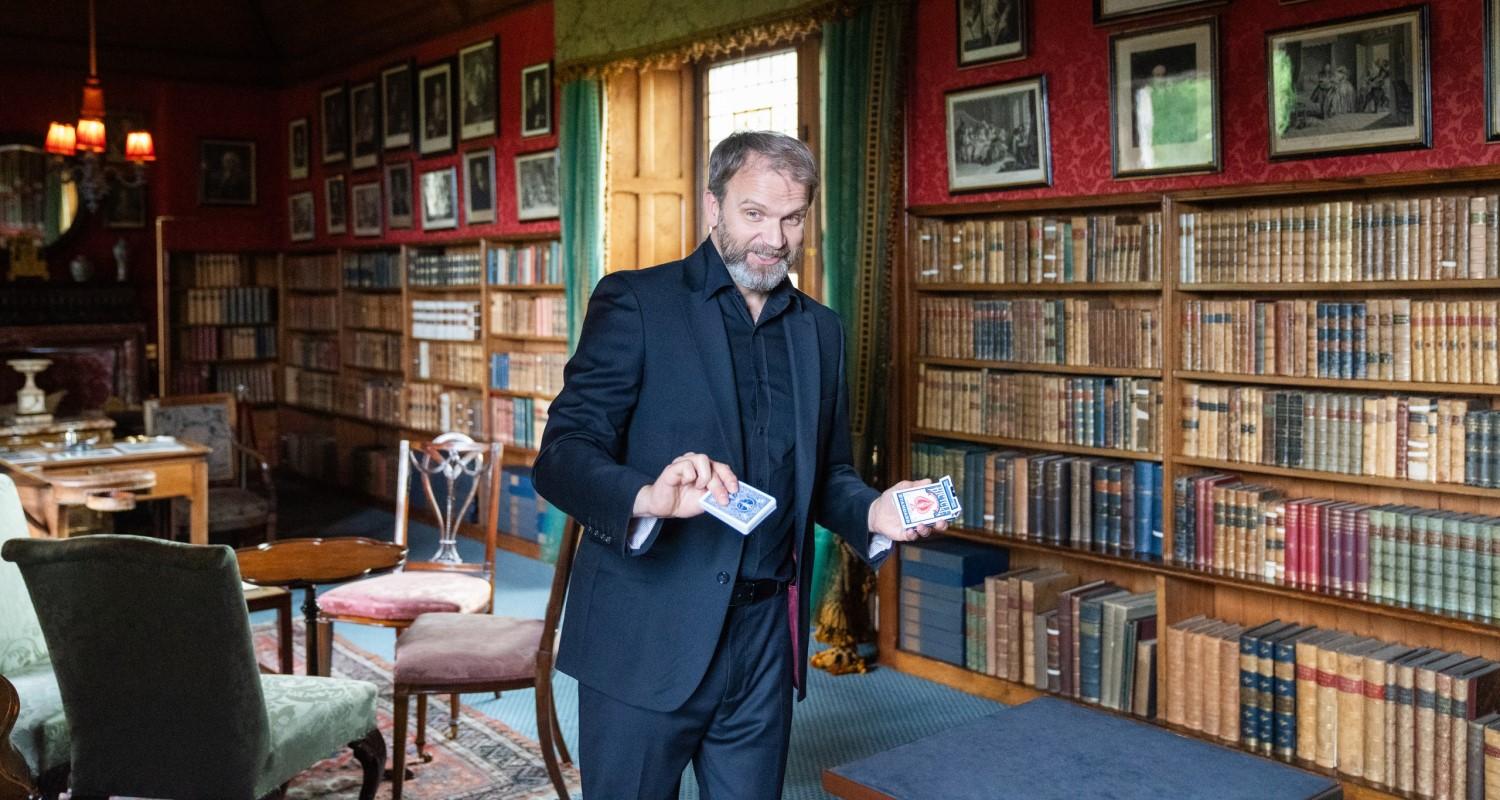 A magician holding a pack of cards standing in the library at Lauriston. The walls are covered in books and pictures.