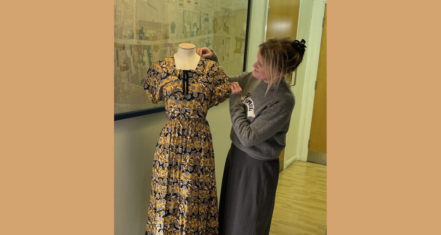 Curator Vicky Garrington, dressed in grey, admiring a yellow and black shortsleeved dress which is displayed on a mannequin. In the background, a street map is on  the wall