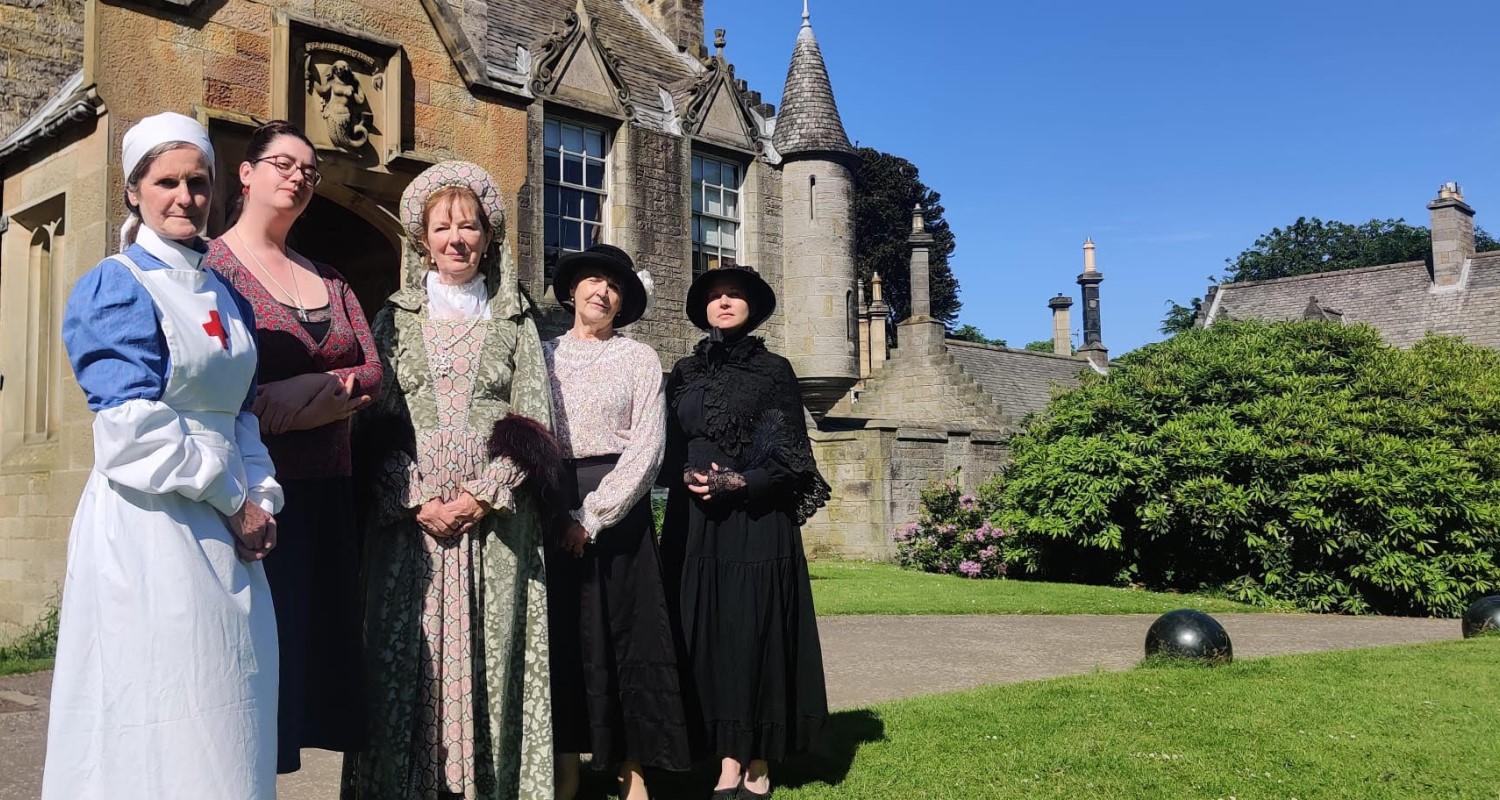 An Edwardian nurse, wealthy lady, and Castle staff stand in front of the entrance in the sunshine