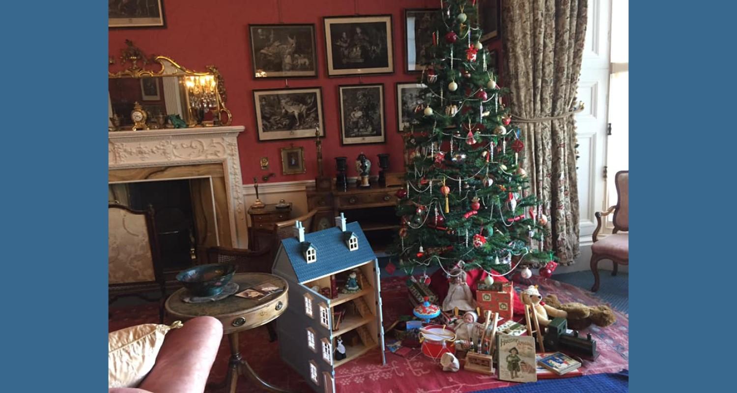 An Edwardian living room with an ornate fireplace, a red Persian rug on a blue carpet, a decorated green Xmas tree with opened gifts underneath, and an open dolls house with a blue roof and furniture inside