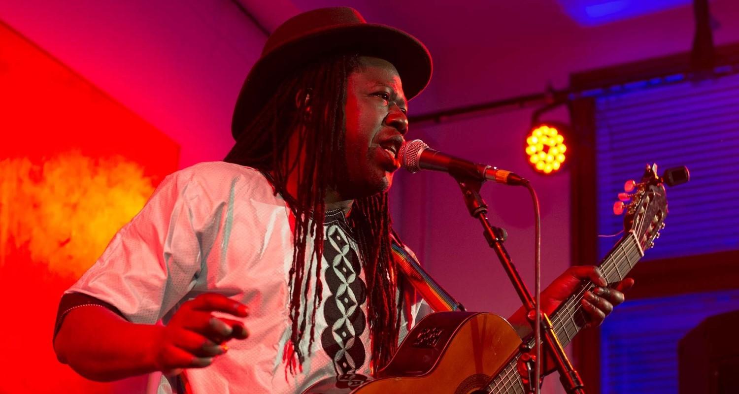 A guy with dreadlocks wearing a trilby and white shirt and holding an acoustic guitar sings into a mic