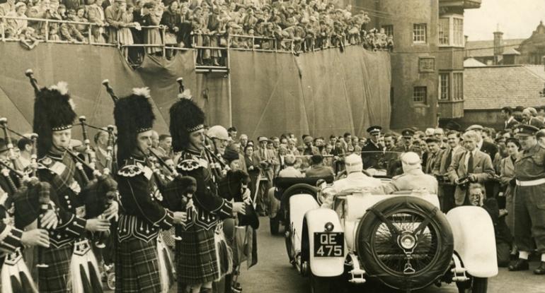 A crowd looking down on two racing cars, each with driver and passenger. Bagpipers stand to the left, more crowds to the right