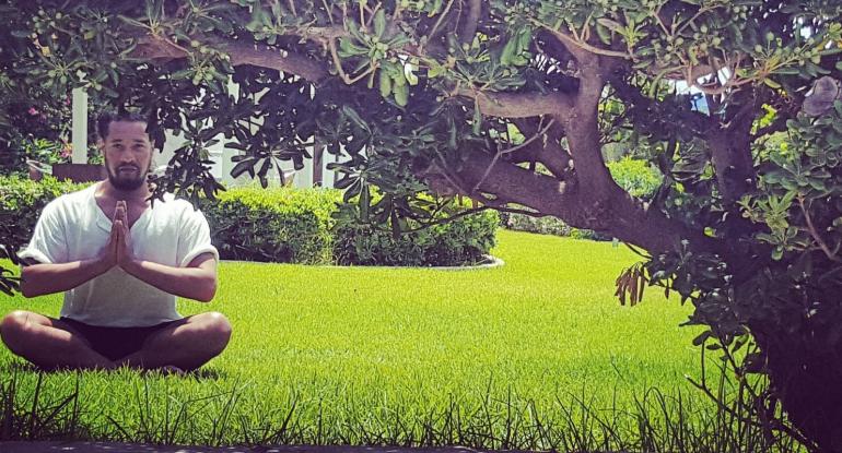 A man in a white tshirt and black shorts sitting in a lotus position on the grass under a tree