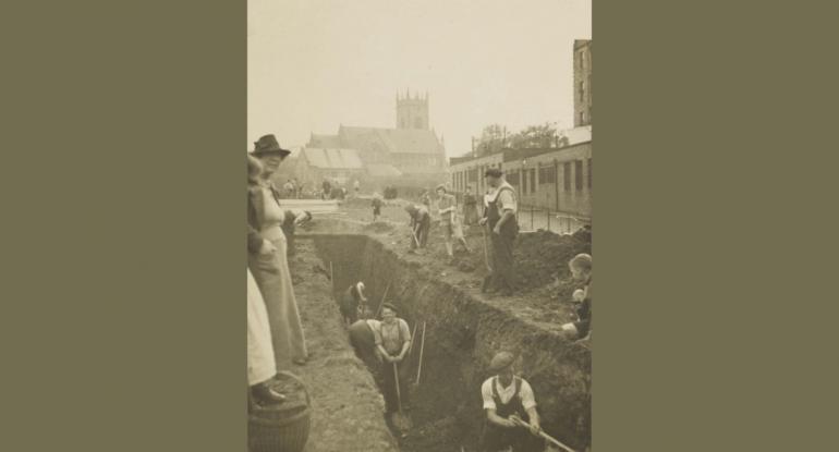 A group of men digging a trench for an air raid shelter in Harrison Park in 1938 watched over by a small boy and two women