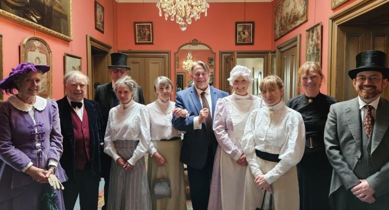 A group of people in Edwardian dress of staff and residents stand in a passageway in the castle beneath a chandelier. The walls are covered in artworks; five doors lead off