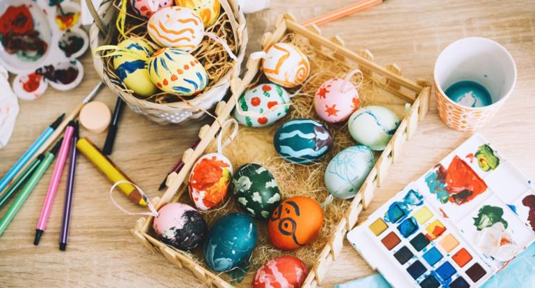 A view of a table top on which rest a box of watercolour paints, baskets of coloured eggs, pencils and a water cup