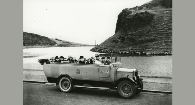 A black and white photo of Charabanc number 502 full of passengers with water and hills in the background