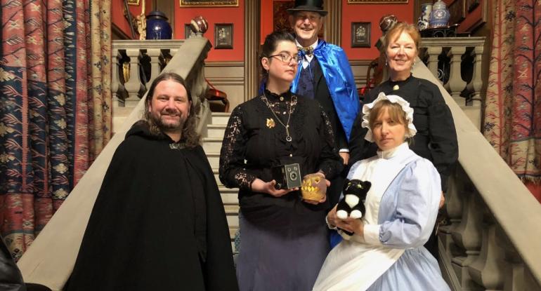 Lauriston Castle stff standing on the staircase, some in cloaks, some in uniform. One holds a turnip lantern, the other a soft toy cat.