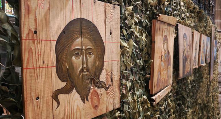 A row of ammo box lids with religious images on them attached to a camouflaged fence