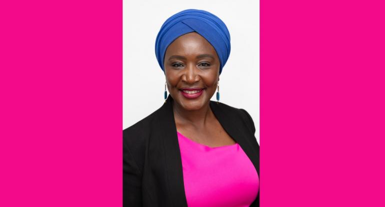 A head and shoulders portrait of Irene Mosota, wearing a pink top, black jacket and blue head wrap