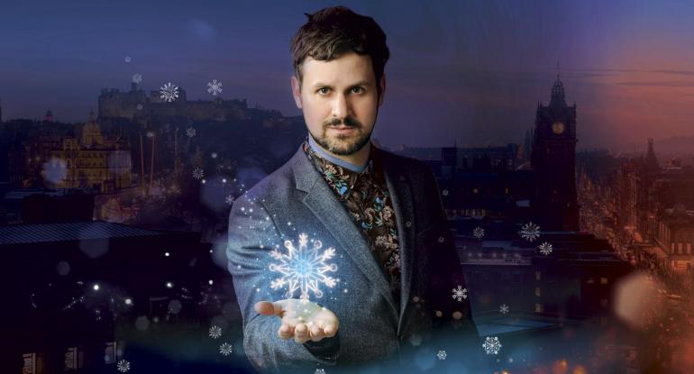 A bearded man stands in front of a dusk backdrop of Edinburgh Castle and Princes Street as a stylised giant snowflake hovers above his outstretched hand