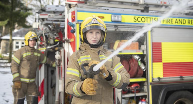 A firefighter in uniform stands in front of a fire engine, hosing an object off-camera