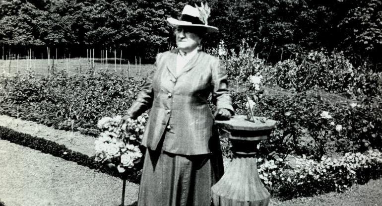 Mrs Reid in a long skirt and jacket, wearing a hat and carrying a cane, standing beside a sundial in the garden. Black and white photo