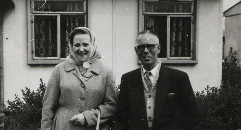 A black and white photo of a middle aged couple standing outside a house. She wears a winter coat and headscarf and has a handbag looped over her arm, he wears a coat over a cardigan, shirt and tie. There is a cigarette in his mouth.