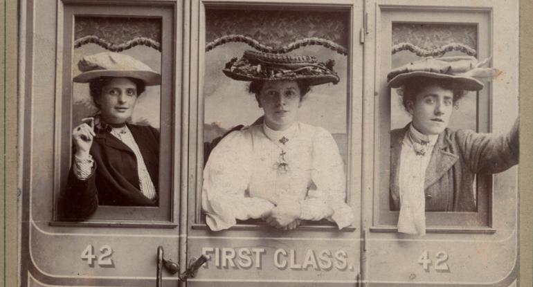 A black and white photo of three women in Edwardian clothes and big hats leaning out of a train window from the first class carriage