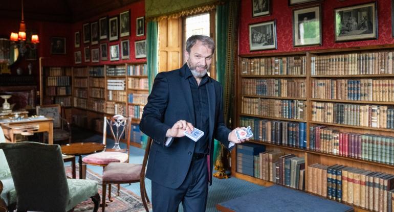 A magician holding a pack of cards standing in the library at Lauriston. The walls are covered in books and pictures.