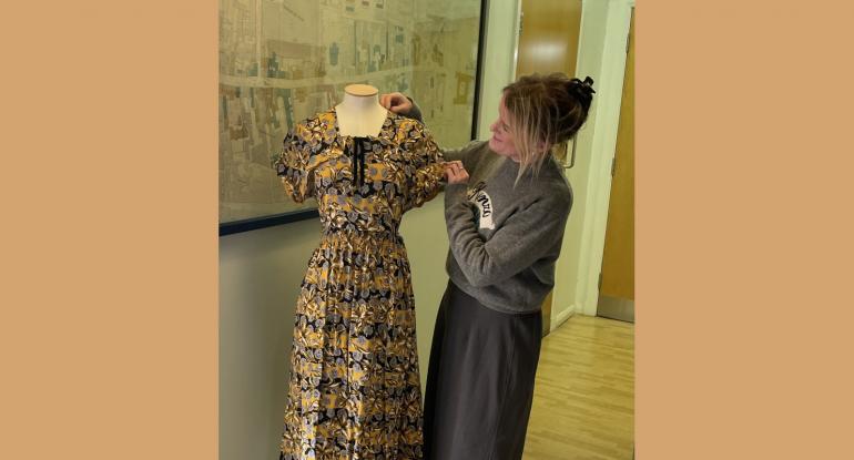 Curator Vicky Garrington, dressed in grey, admiring a yellow and black shortsleeved dress which is displayed on a mannequin. In the background, a street map is on  the wall