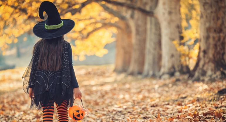 A child dressed as a witch with hat, black cape and black and orange tights, carrying a plastic pumpkin, and walking through fallen autumn leaves