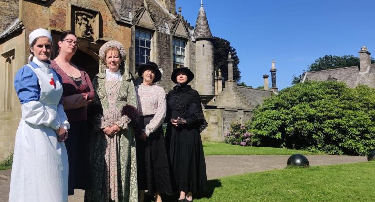 An Edwardian nurse, wealthy lady, and Castle staff stand in front of the entrance in the sunshine