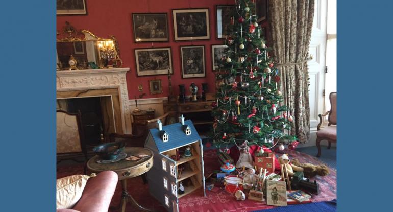 An Edwardian living room with an ornate fireplace, a red Persian rug on a blue carpet, a decorated green Xmas tree with opened gifts underneath, and an open dolls house with a blue roof and furniture inside