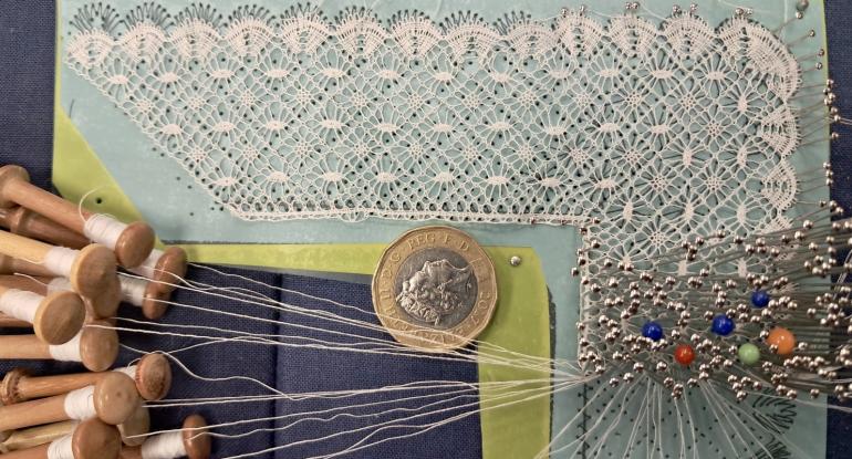 Lacework in progress with pins and bobbins and a £2 coin for scale